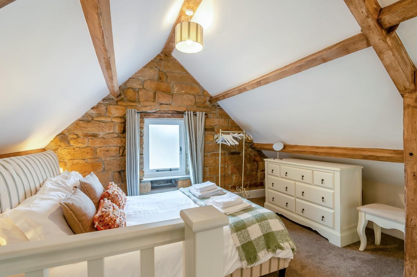 A bedroom with a stone wall, a bed with white bedding, a dresser, and a window with curtains.