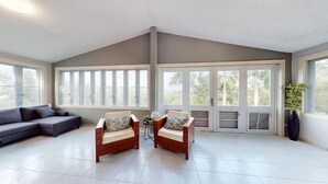 Amplia sala de estar con un sofá seccional gris, dos sillones de madera con cojines, una mesita con una planta y ventanas amplias que dan a zonas verdes.