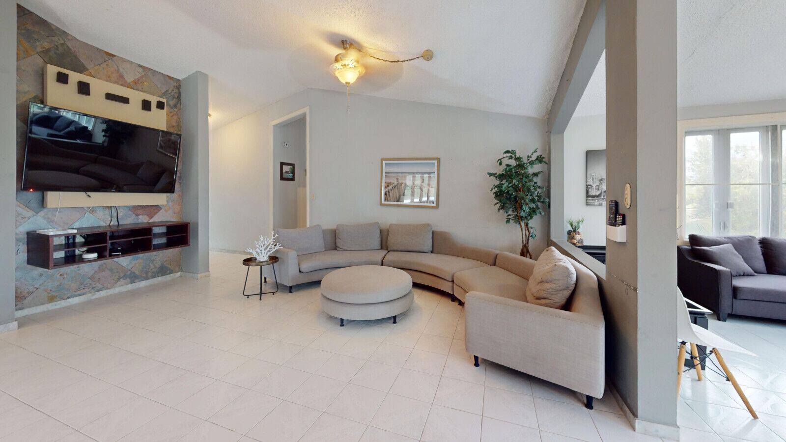 A spacious living room with a large L-shaped sectional sofa, a round ottoman, and a flat-screen TV mounted on a stone wall.