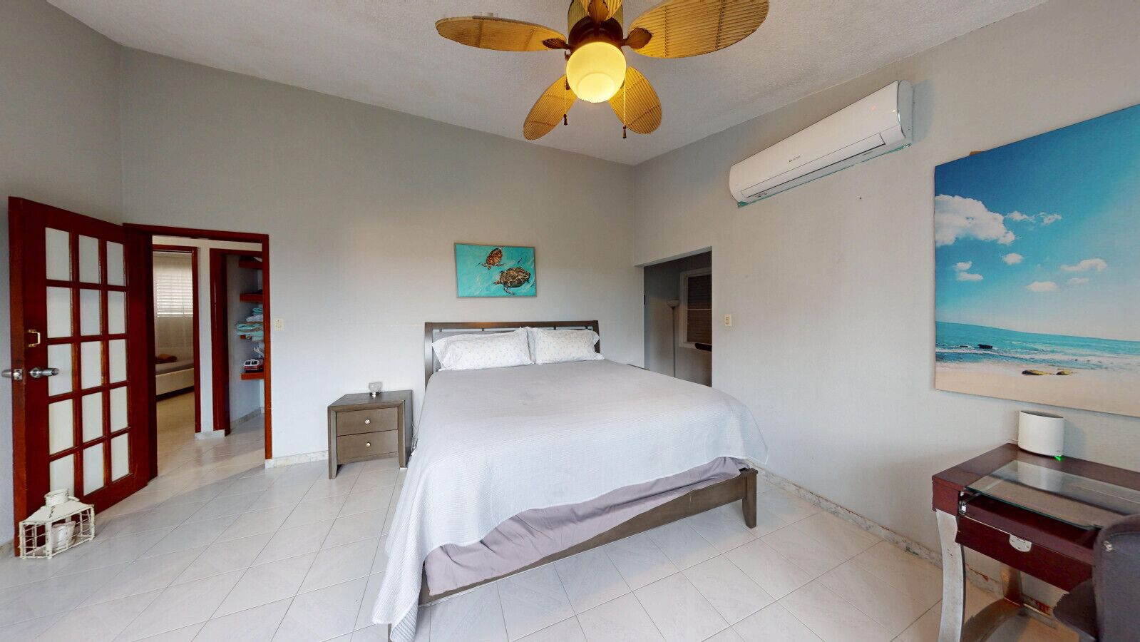 Un dormitorio con cama, mesita de noche, escritorio, ventilador de techo, aire acondicionado, un cuadro y una puerta que da a otra habitación.