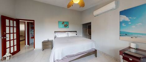 Un dormitorio con cama, mesita de noche, escritorio, ventilador de techo, aire acondicionado, un cuadro y una puerta que da a otra habitación.