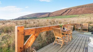 Um deck de madeira com cadeiras e uma mesa, com vista para um campo gramado e colinas.