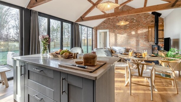 Een moderne keuken met een groot eiland, houten balken, een bakstenen muur en uitzicht op het omliggende landschap.