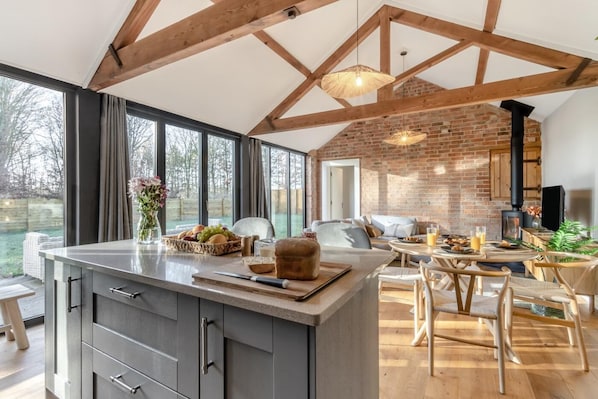 A modern kitchen with a large island, wooden beams, a brick wall, and a view of the outdoors. - 2 Bed in Market Harborough (oc-o34219) (Clipston)