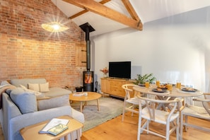 A cozy living room with a brick wall, a fireplace, a sofa, a TV, and a dining area with chairs and a table. - 2 Bed in Market Harborough (oc-o34219) (Clipston)