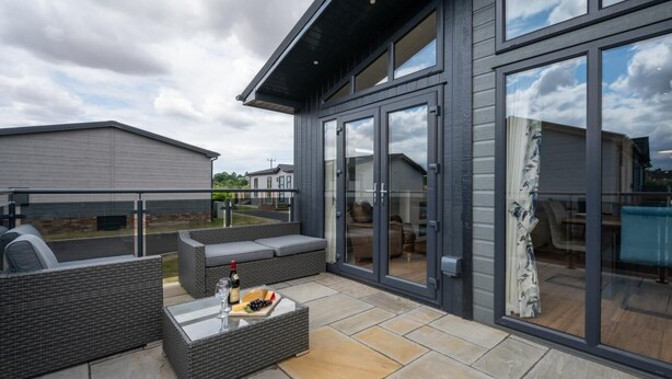 A modern outdoor patio with a glass-enclosed seating area, a glass-top table with a bottle and snacks, and a view of the surrounding neighborhood. - 3 Bed in Burnham Market (oc-2334) (Burnham Market)