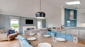A modern living room with a dining table set for six, a sofa, and a kitchen area with a blue accent wall. - 3 Bed in Burnham Market (oc-2334) (Burnham Market)