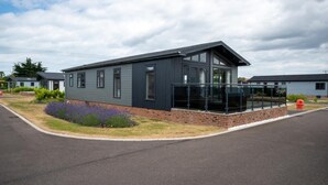 A modern single-story house with a dark exterior, a brick foundation, and a small front yard with purple flowers. - 3 Bed in Burnham Market (oc-2334) (Burnham Market)