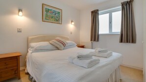 A neatly made bed with pillows and folded towels, a bedside table, a nightstand, a window with curtains, and a wall-mounted light fixture.