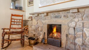Una chimenea de piedra con fuego, un sillón mecedor de madera, un mapa en la pared y un estante con una radio.
