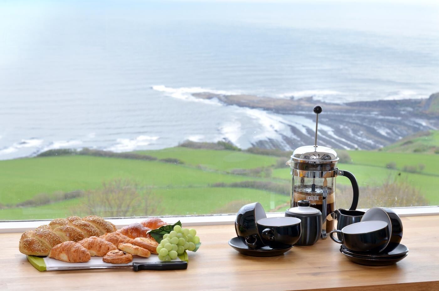 Desayuno con cruasanes, uvas y una cafetera sobre una mesa con vistas panorámicas al océano.