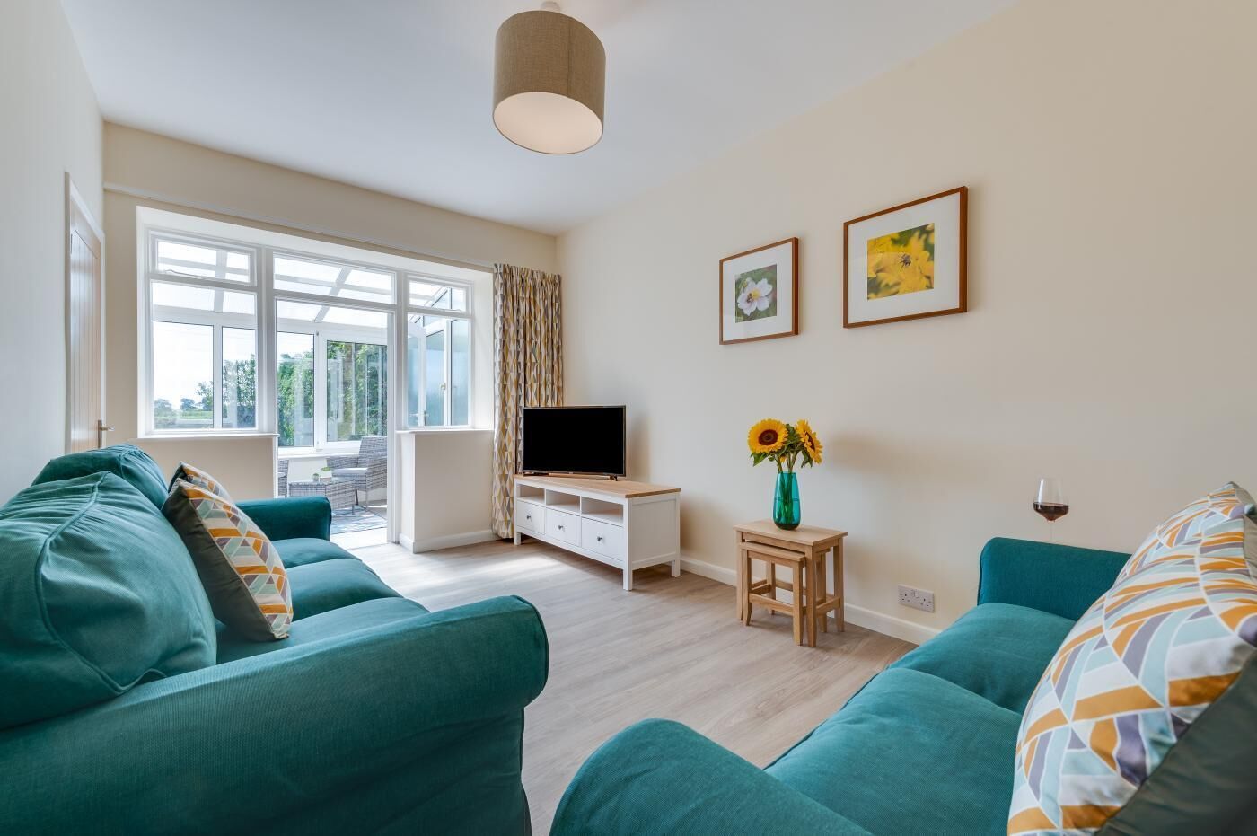 A modern living room with teal sofas, a TV on a white cabinet, a small wooden table with a vase of sunflowers, and two framed pictures on the wall.