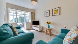 A modern living room with teal sofas, a TV on a white cabinet, a small wooden table with a vase of sunflowers, and two framed pictures on the wall. - 2 Bed in Alnwick (oc-nb1986) (Howick)