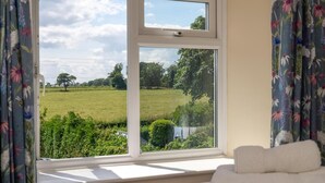 A room with a large window overlooking a green field and trees, a white towel on a chair, and floral curtains. - 2 Bed in Alnwick (oc-nb1986) (Howick)