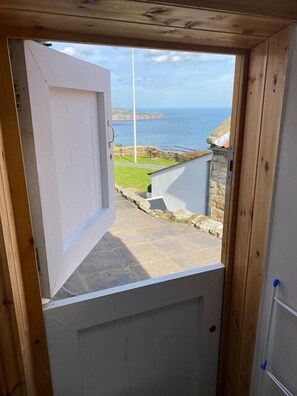 Blick aus einem Zimmer mit einer Holztür, die auf einen Balkon mit Blick auf eine Küstenlandschaft mit Steinmauern und einer Rasenfläche führt.