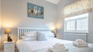 A bedroom with a bed, white nightstands, a lamp, a painting, and a window with shutters.