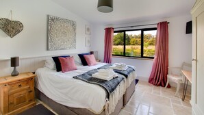 Un dormitorio con cama, mesita de noche, lámpara, una obra de arte, ventana con cortinas y vista a zonas verdes.