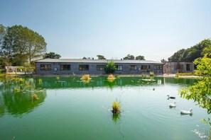 A rustic cabin with a pond in front, surrounded by trees and greenery. - 5 Bed in Briston (oc-1875) (Briston)