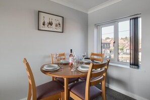 Un coin repas avec une table ronde dressée pour quatre personnes, des chaises en bois, un tableau encadré au mur et une fenêtre donnant sur des immeubles.