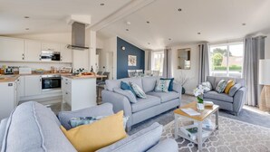 A modern living room with a grey sofa, a coffee table, and a kitchen area with white cabinets and a built-in oven. - 2 Bed in Burnham Market (oc-2335) (Burnham Market)