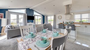 A modern dining area with a table set for four, a kitchen in the background, and a large window providing natural light. - 2 Bed in Burnham Market (oc-2335) (Burnham Market)