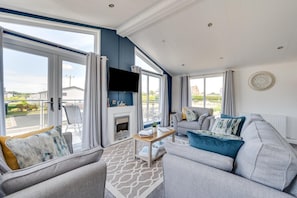 A modern living room with a fireplace, comfortable seating, and large windows offering a view of the outdoors. - 2 Bed in Burnham Market (oc-2335) (Burnham Market)