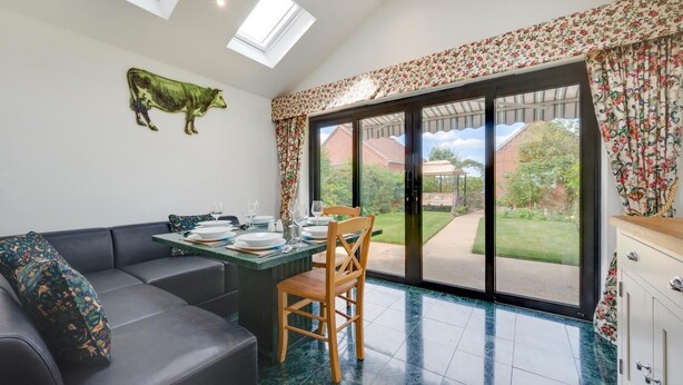 A dining area with a table set for four, a sofa, and a large window with a view of a garden. - 3 Bed in Holt (oc-2280) (Holt)