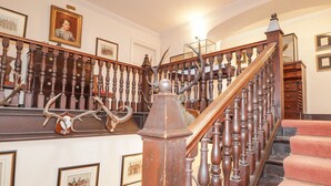 Een statige trap met een houten balustrade, decoraties van gewei en ingelijste historische schilderijen aan de muren.