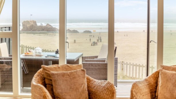 Une vue sur la plage depuis une chambre avec une table et des chaises, une clôture en bois et une formation rocheuse au loin.