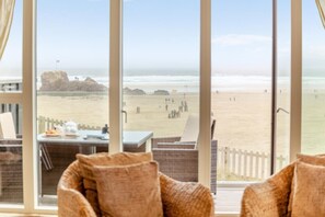 Vista a la playa desde una habitación con una mesa y sillas, una cerca de madera y una formación rocosa a lo lejos.