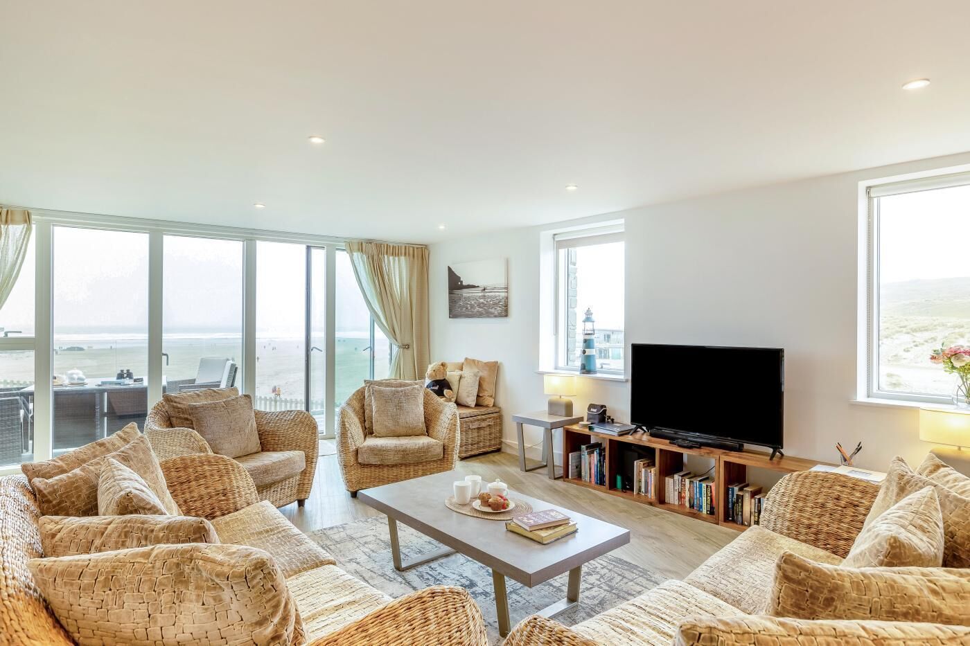 A spacious living room with wicker furniture, a flat-screen TV, and a bookshelf.