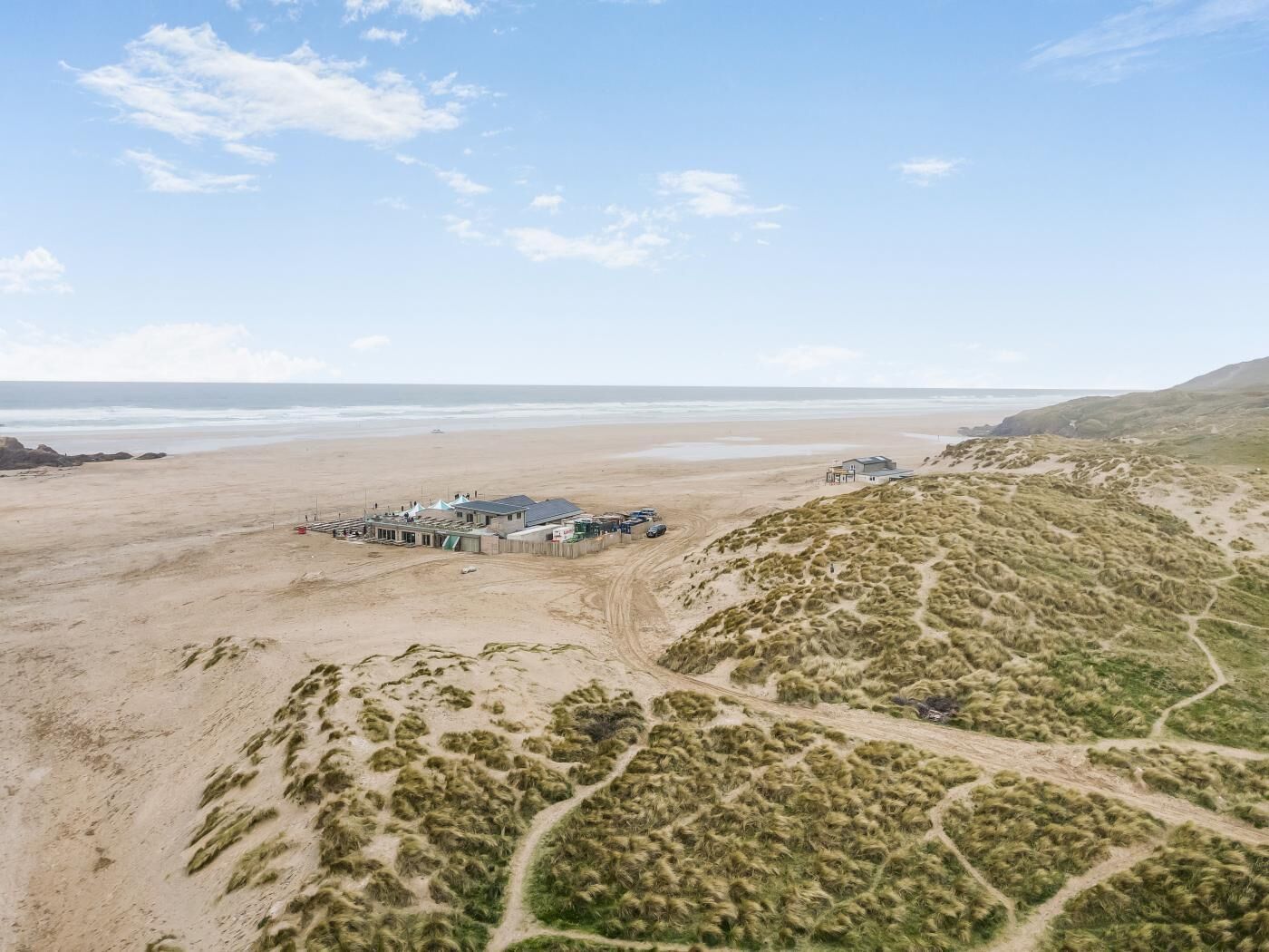 An aerial view of a coastal area with a building complex near the beach, sandy dunes, and a clear sky.