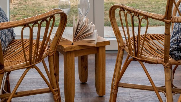 A room with two wicker chairs and a small wooden table, a door leading to an outdoor area with a fence and hay bales, and a view of the sky.