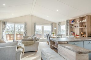 Un salon spacieux avec une grande fenêtre, un coin repas et une cuisine dotée d’étagères en bois.