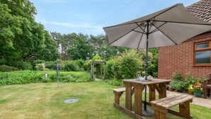 Un espace patio avec une table et des bancs en bois, un grand parasol et une maison en briques en arrière-plan.