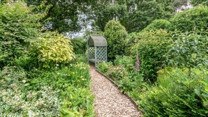 Un sentier de jardin mène à un belvédère en bois, entouré d’une végétation luxuriante et de diverses plantes à fleurs.