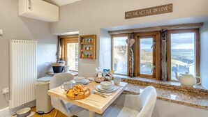 A cozy dining area with a wooden table set for two, a view of the countryside through large windows, and a sign above the windows that reads "GET THE KETTLE ON LOVE". - 1 Bed in Slaithwaite (oc-c34249) (Ripponden)