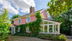 Uma casa de dois andares com fachada rosa, uma estufa com molduras brancas e um caminho de acesso de cascalho.