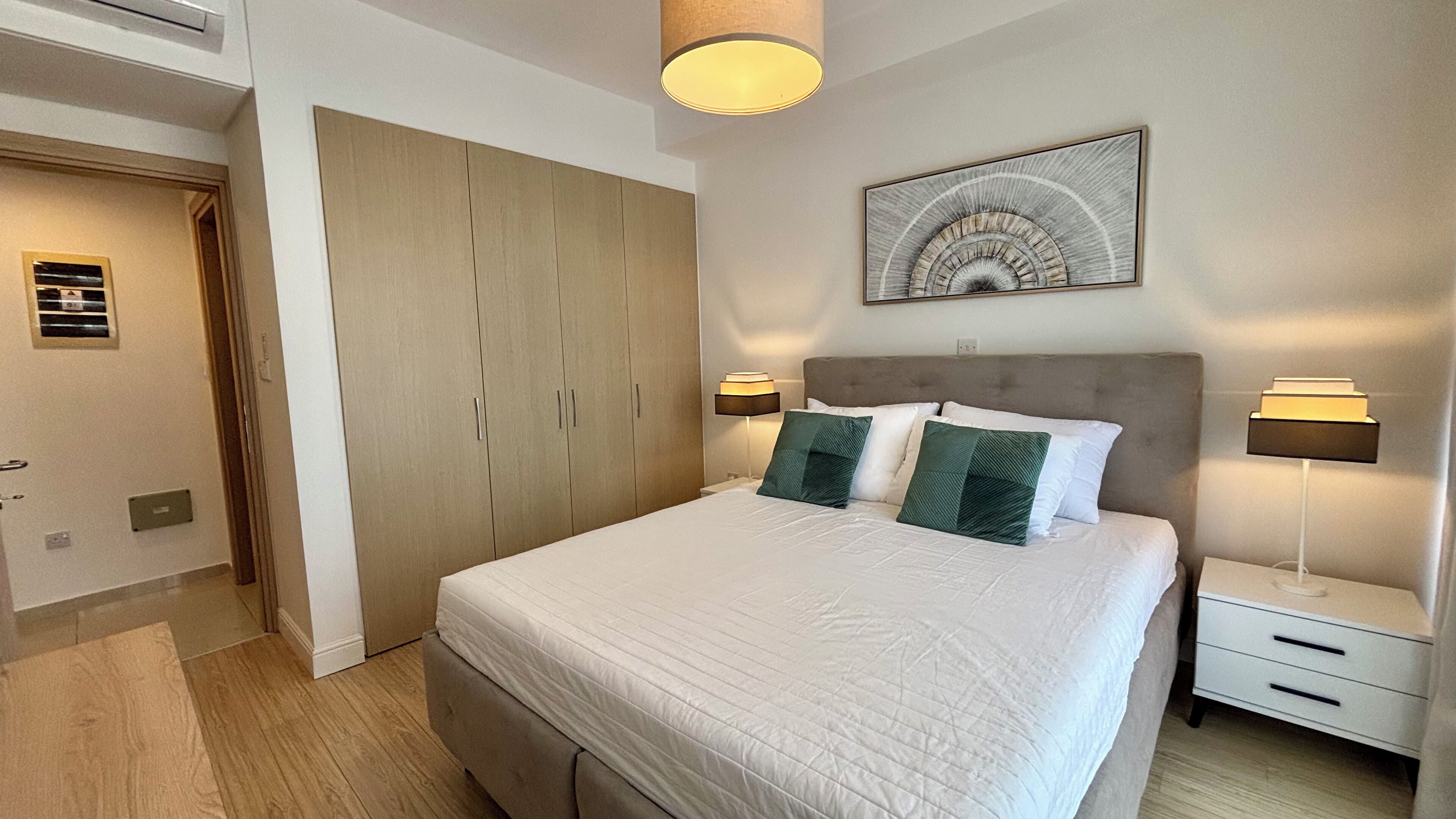 A bedroom with a large bed, two bedside tables, a headboard, a wardrobe, and a ceiling light fixture.