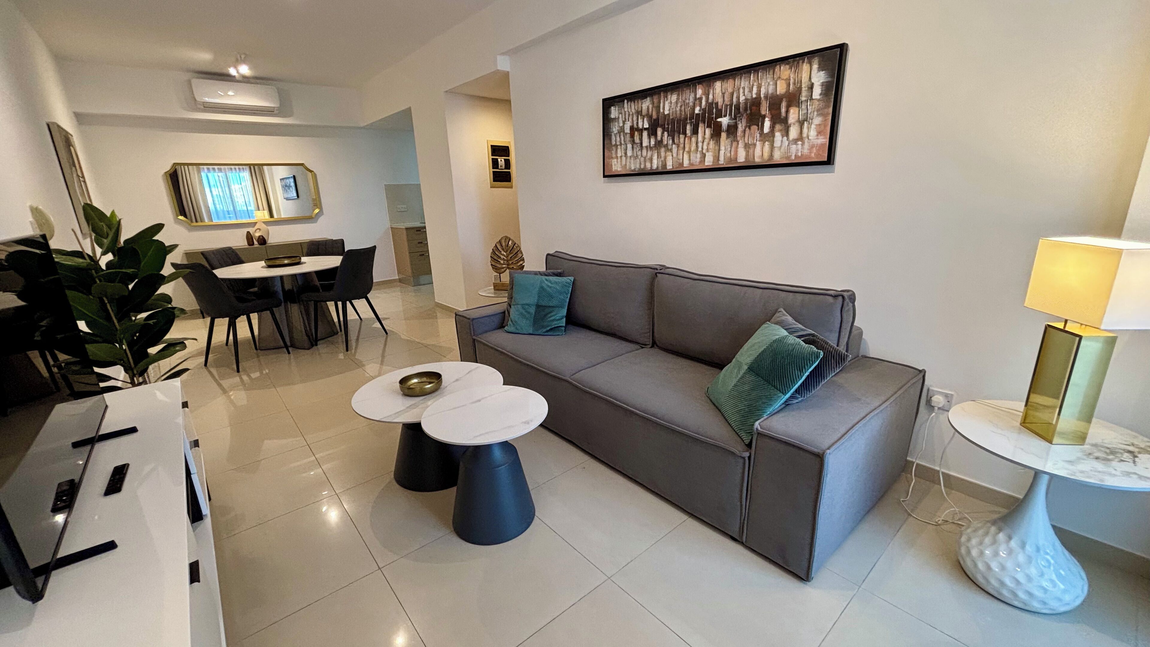 A modern living room with a grey sofa, two white round tables, and a small side table with a lamp. There is a dining area with a table and chairs in the background.