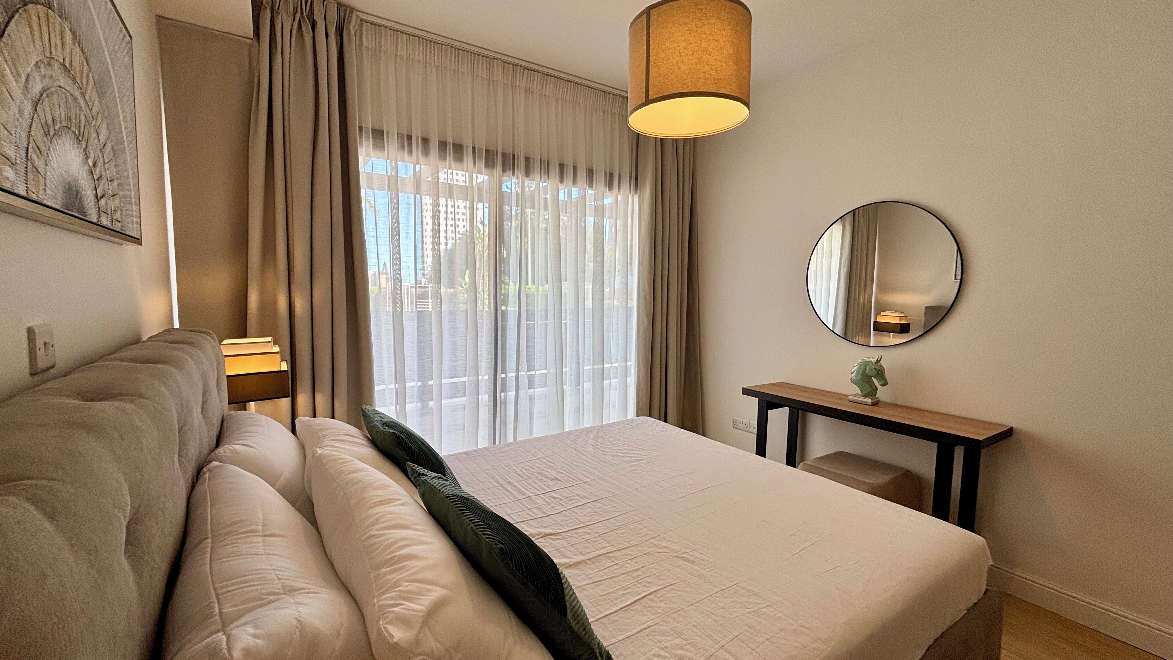Une chambre d’hôtel avec un lit, une table de chevet avec une lampe, un miroir et une vue sur l’extérieur à travers des rideaux transparents.