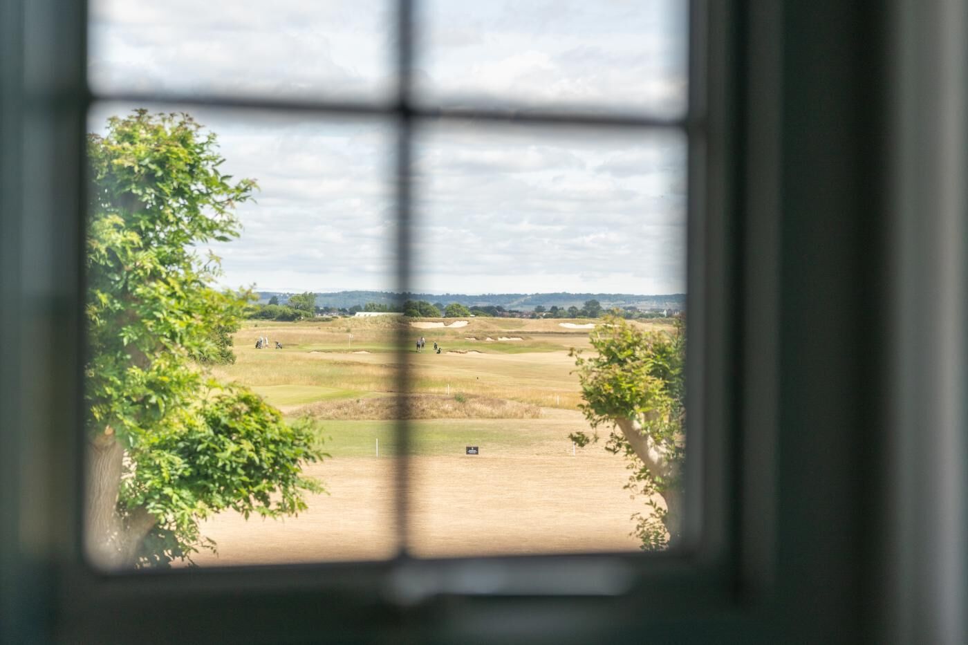 Vista pela janela com padrão quadriculado, mostrando um campo de golfe com árvores e céu claro.