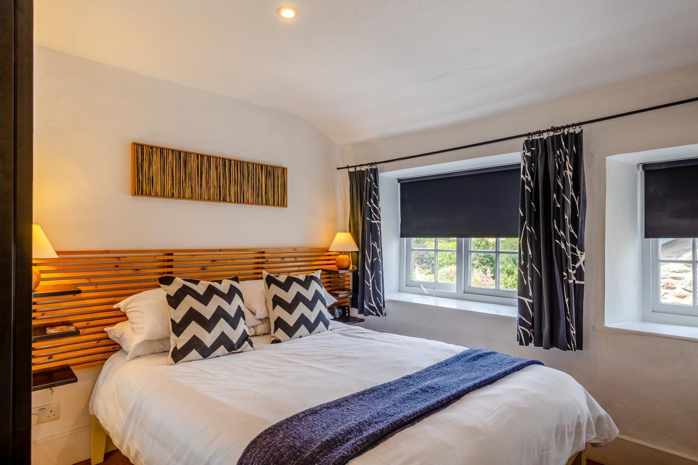 A bedroom with a bed, two bedside tables, a wall-mounted artwork, a window with curtains, and a view of greenery outside.