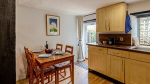 A dining area with a wooden table set for four, a kitchen with wooden cabinets, and a window with a curtain. - 3 Bed in Eype (oc-e34183) (Eype)