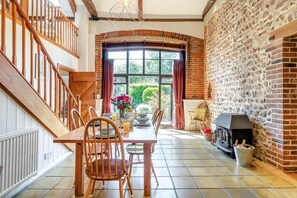 A dining area with a brick fireplace, wooden staircase, and a view of greenery outside.
