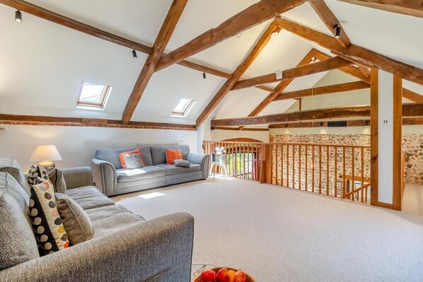 A spacious loft with exposed wooden beams, a grey sofa with orange cushions, and a view of the outside through a skylight.