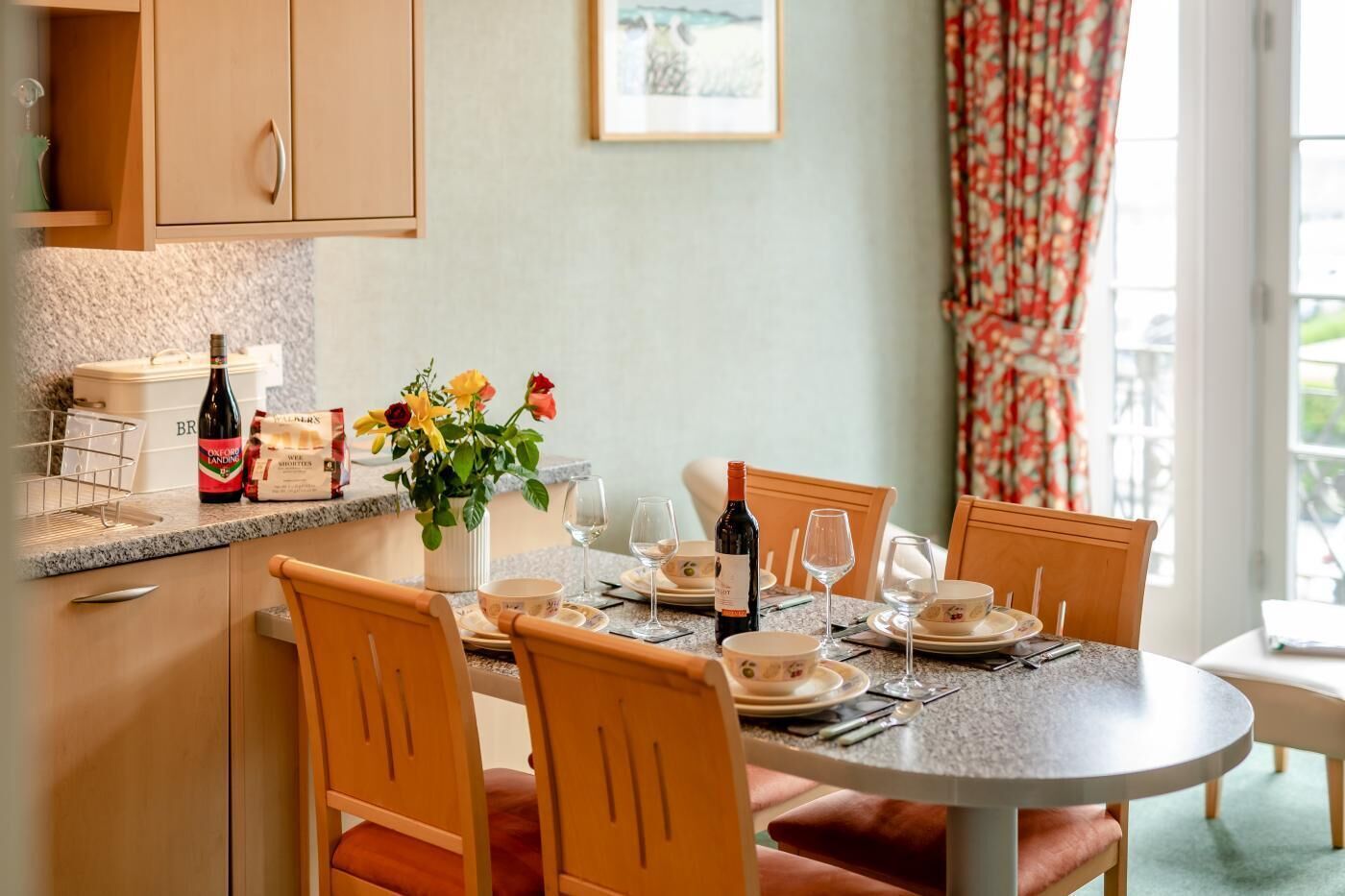 A dining area with a round table set for four, wooden chairs, a vase of flowers, and a bottle of wine on the counter.