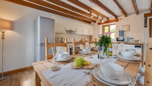 A well-lit kitchen with wooden beams, a dining table set for four with white plates and glasses, and a refrigerator. - 3 Bed in Aberaeron (oc-bow315) (Bethania)