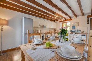 Eine helle Küche mit Holzbalken, einem Esstisch für vier Personen, weißen Tellern und Gläsern sowie einem Kühlschrank.