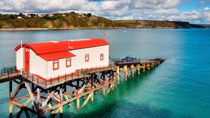 A red and white building on a pier extending into a calm sea, with a boat docked nearby. - 2 Bed in Tenby (oc-pw1031) (Tenby)